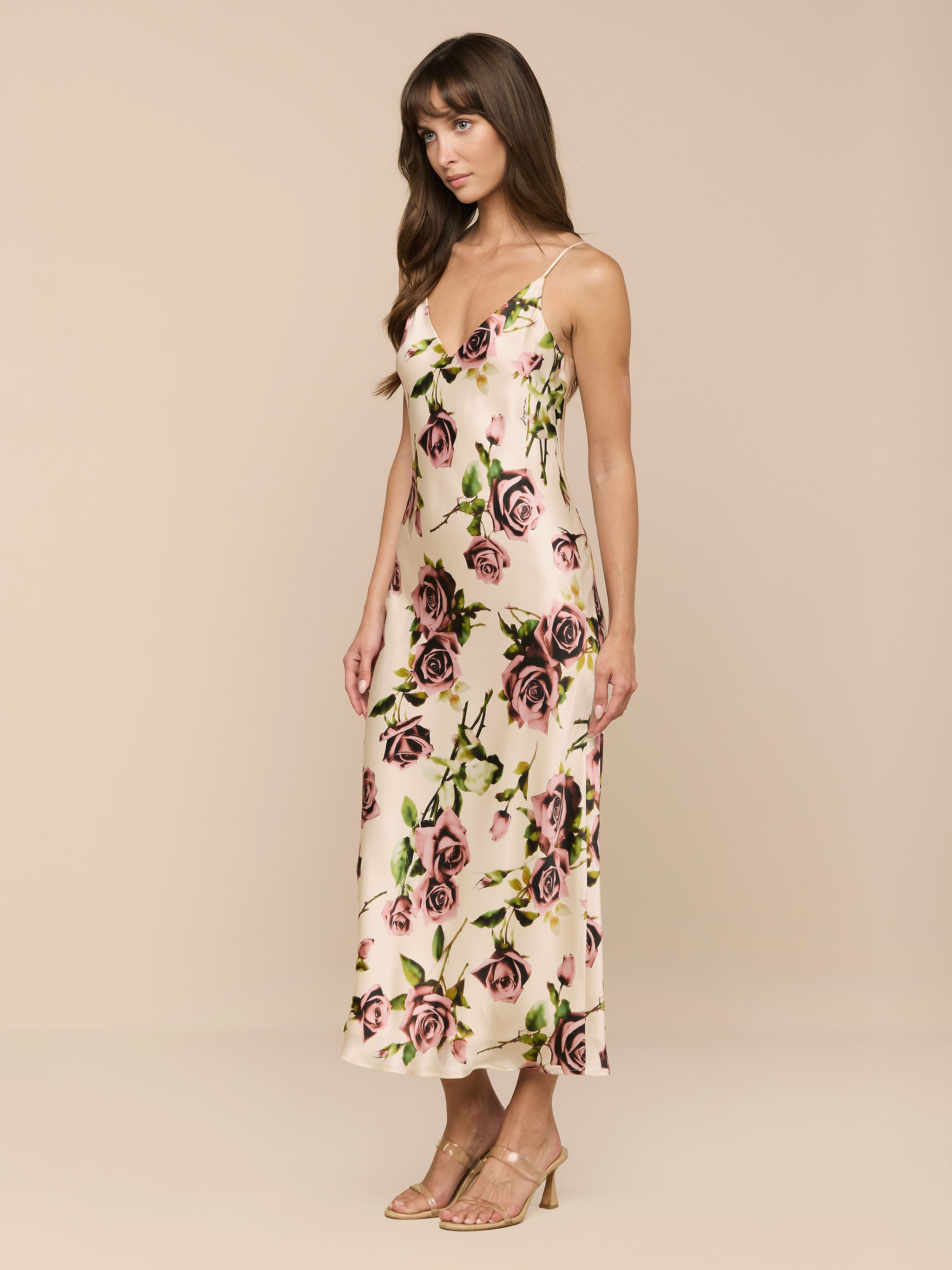Una mujer lleva el vestido Seridie Silk Slip Dress en rosa claro con un delicado estampado de rosas, cortado al bies y con tirantes espagueti, combinado con sandalias de tacón nude, de lado sobre un fondo beige liso.