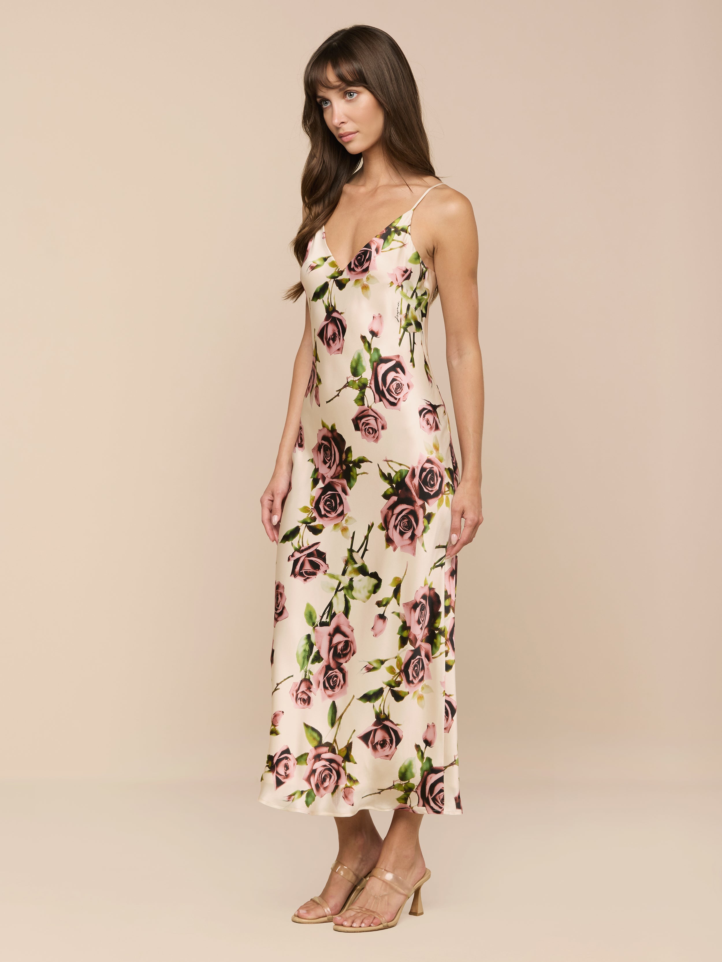 Una mujer lleva el vestido Seridie Silk Slip Dress en rosa claro con un delicado estampado de rosas, cortado al bies y con tirantes espagueti, combinado con sandalias de tacón nude, de lado sobre un fondo beige liso.
