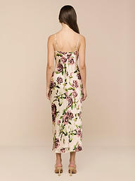 Una mujer con el pelo largo y castaño lleva el vestido Seridie Silk Slip Dress, con corte al bies de color marfil y estampado de rosas de color rosa verdoso. Lo combina con sandalias de tacón beige, sobre un fondo beige liso y de espaldas a la cámara.