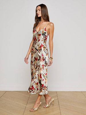 A woman with long brown hair poses indoors against a white wall, wearing the Seridie Silk Slip Dress—an ankle-length charmeuse slip featuring a colorful abstract floral and newspaper print—and heeled sandals.