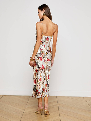 A woman with long brown hair wears the Seridie Silk Slip Dress, a fitted silk midi dress with thin straps and colorful floral and newspaper print, paired with large earrings and clear high heels on a wooden floor.