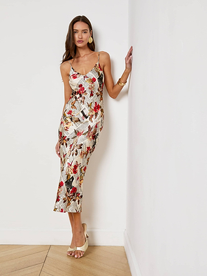 A woman in the Seridie Silk Slip Dress—a sleeveless, ankle-length slip with spaghetti straps—stands against a white wall, wearing beige heels and gold earrings, with one hand touching the wall.