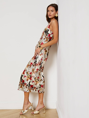 A woman in the Seridie Silk Slip Dress, featuring a colorful newspaper print and sleeveless midi silhouette, leans against a white wall. She completes her look with gold hoop earrings, a bracelet, open-toe heels, and minimal accessories.