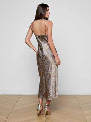 A woman with long brown hair stands facing away, wearing the Seridie Silk Slip Dress, a shiny paisley print slip dress with thin straps, paired with metallic high-heeled sandals on a light wood floor against a white wall.