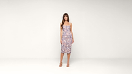 A woman stands in a simple studio wearing a fitted, sleeveless, knee-length dress with a multicolored pattern and strappy sandals. The background is plain and light-colored.