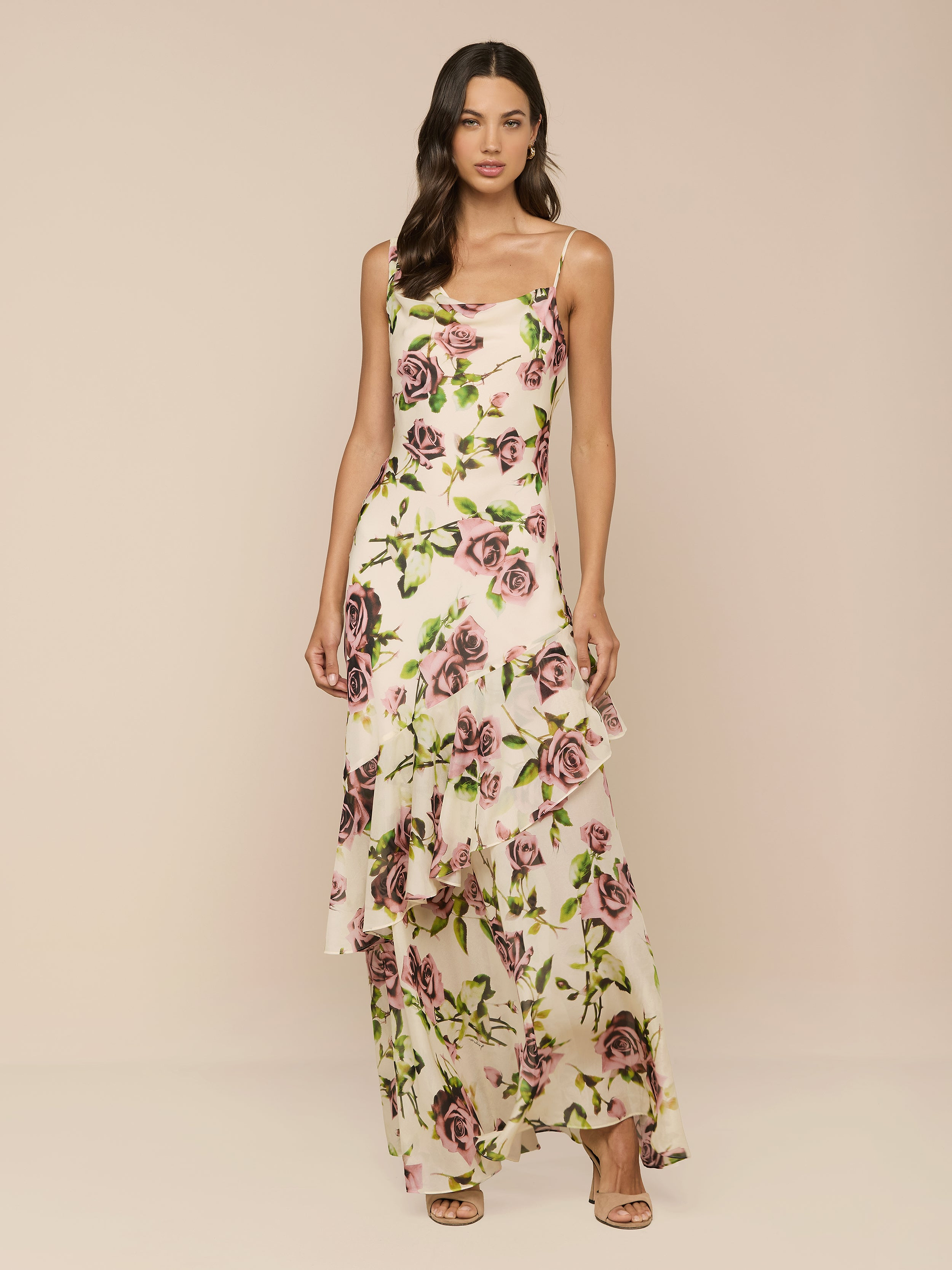 A woman with long, wavy brown hair wears the Viola Silk Cowl-Neck Gown, featuring a sleeveless design, mauve rose print, ruffled hem, and thin straps. She pairs it with beige open-toe heels against a neutral background.