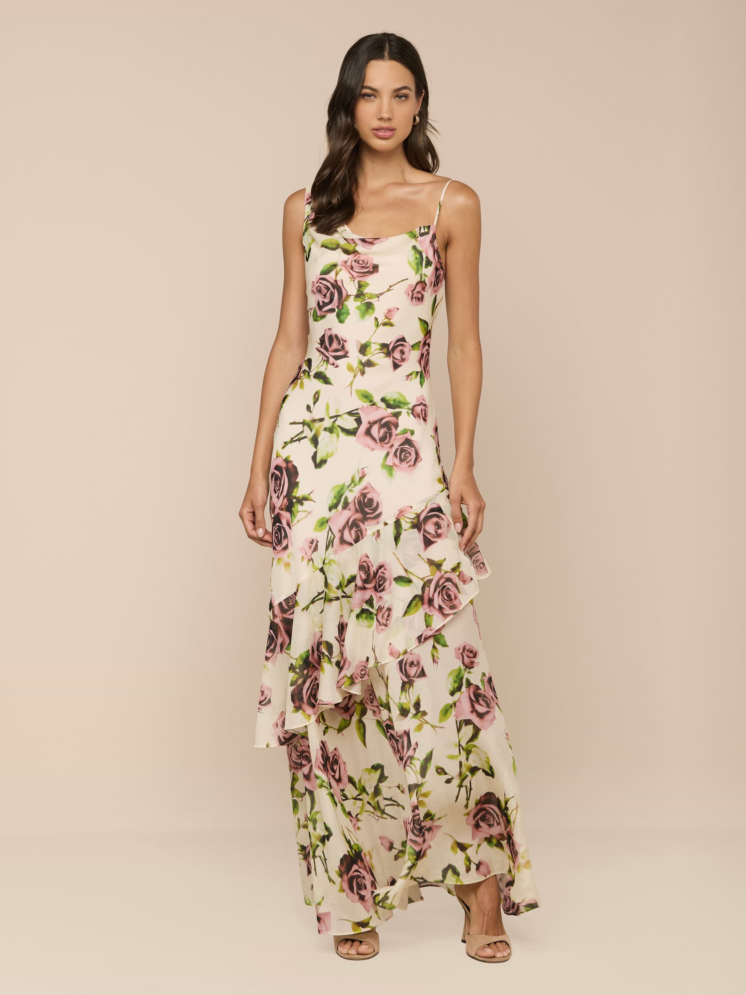 A woman with long, wavy brown hair wears the Viola Silk Cowl-Neck Gown, featuring a sleeveless design, mauve rose print, ruffled hem, and thin straps. She pairs it with beige open-toe heels against a neutral background.