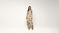 Woman standing in a light-colored, sleeveless floral maxi dress and sandals against a plain white background.
