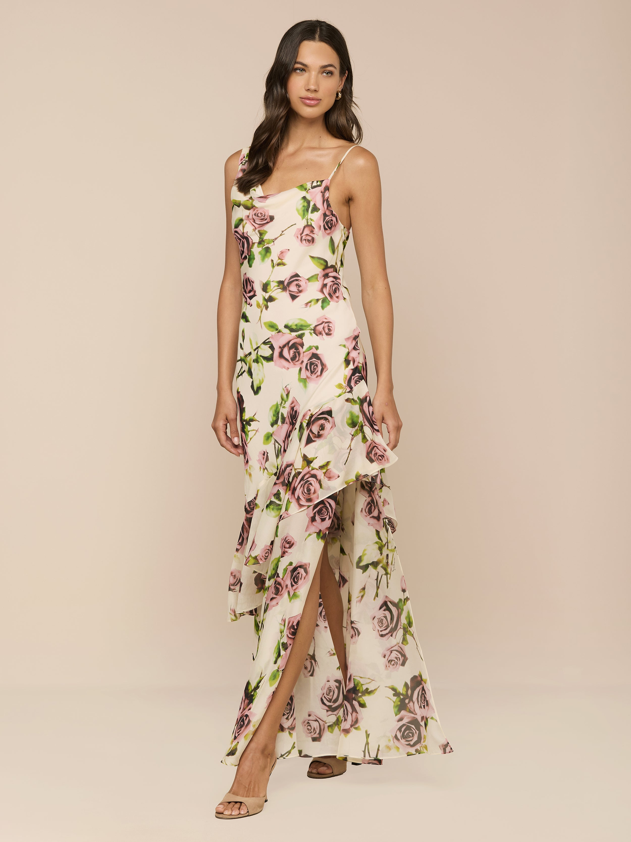 A woman poses against a beige background in the Viola Silk Cowl-Neck Gown, showcasing its mauve rose print, sleeveless design, asymmetrical ruffled hem, thigh-high slit, strappy sandals, and wavy hair.