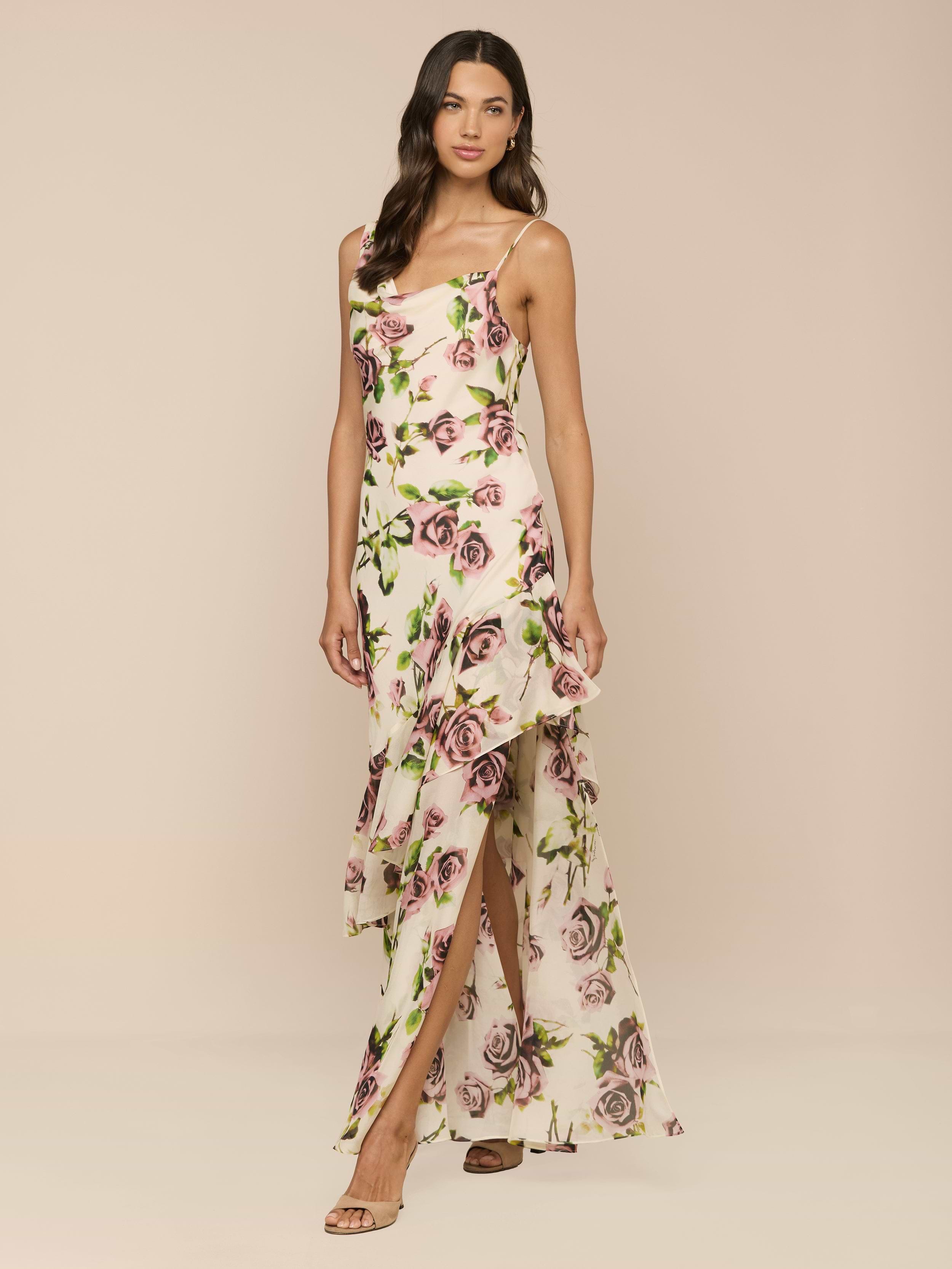 A woman poses against a beige background in the Viola Silk Cowl-Neck Gown, showcasing its mauve rose print, sleeveless design, asymmetrical ruffled hem, thigh-high slit, strappy sandals, and wavy hair.