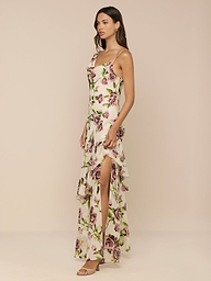 A woman poses against a neutral background in the Viola Silk Cowl-Neck Gown, featuring a light mauve rose print, high slit, ruffled layers, sleeveless design, and strappy sandals. She has long dark hair.