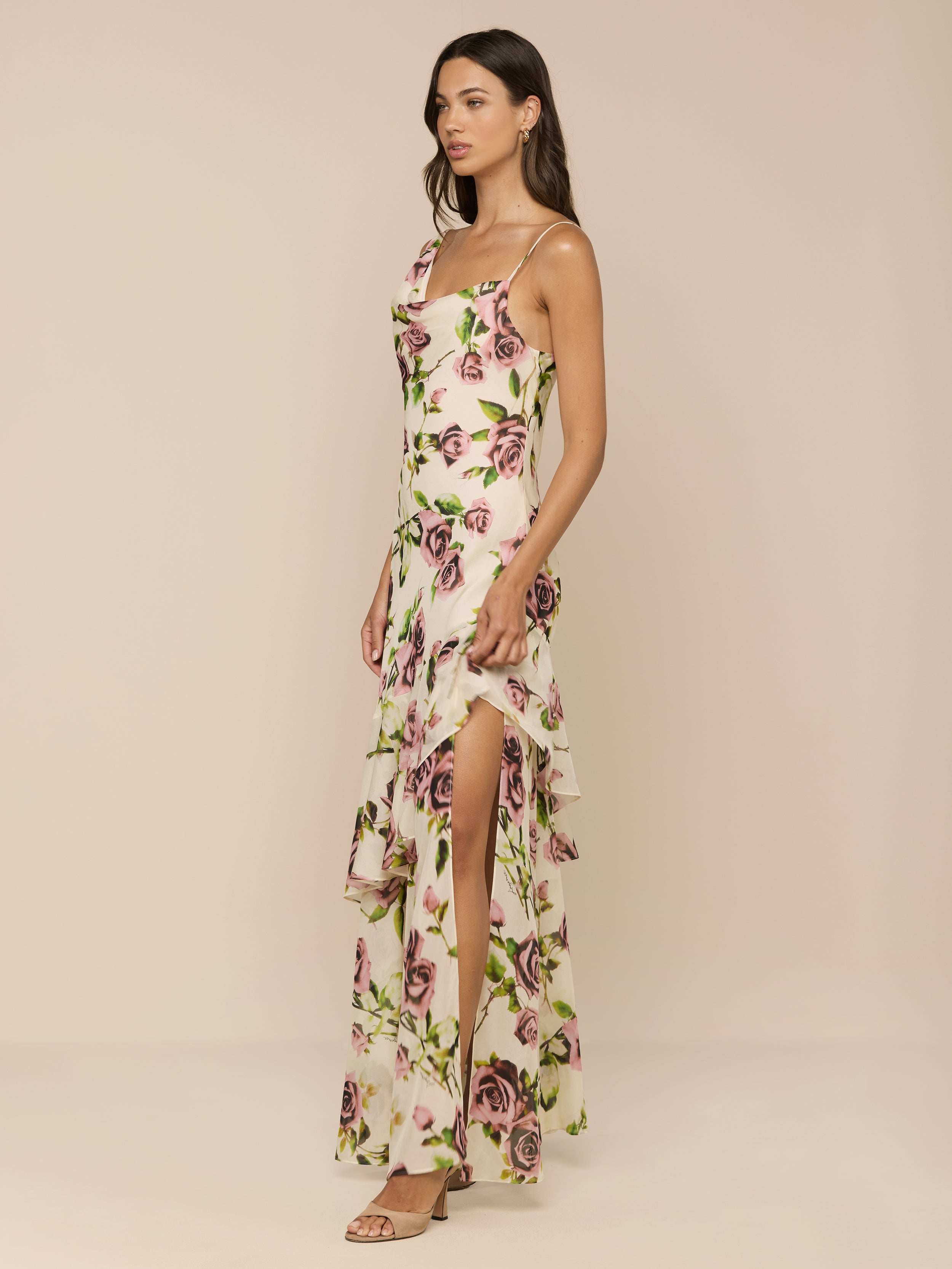 A woman poses against a neutral background in the Viola Silk Cowl-Neck Gown, featuring a light mauve rose print, high slit, ruffled layers, sleeveless design, and strappy sandals. She has long dark hair.