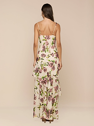 A woman stands facing away, wearing the Viola Silk Cowl-Neck Gown—an ankle-length dress in cream silk chiffon with a mauve rose print, thin straps, and layered asymmetric ruffle details.