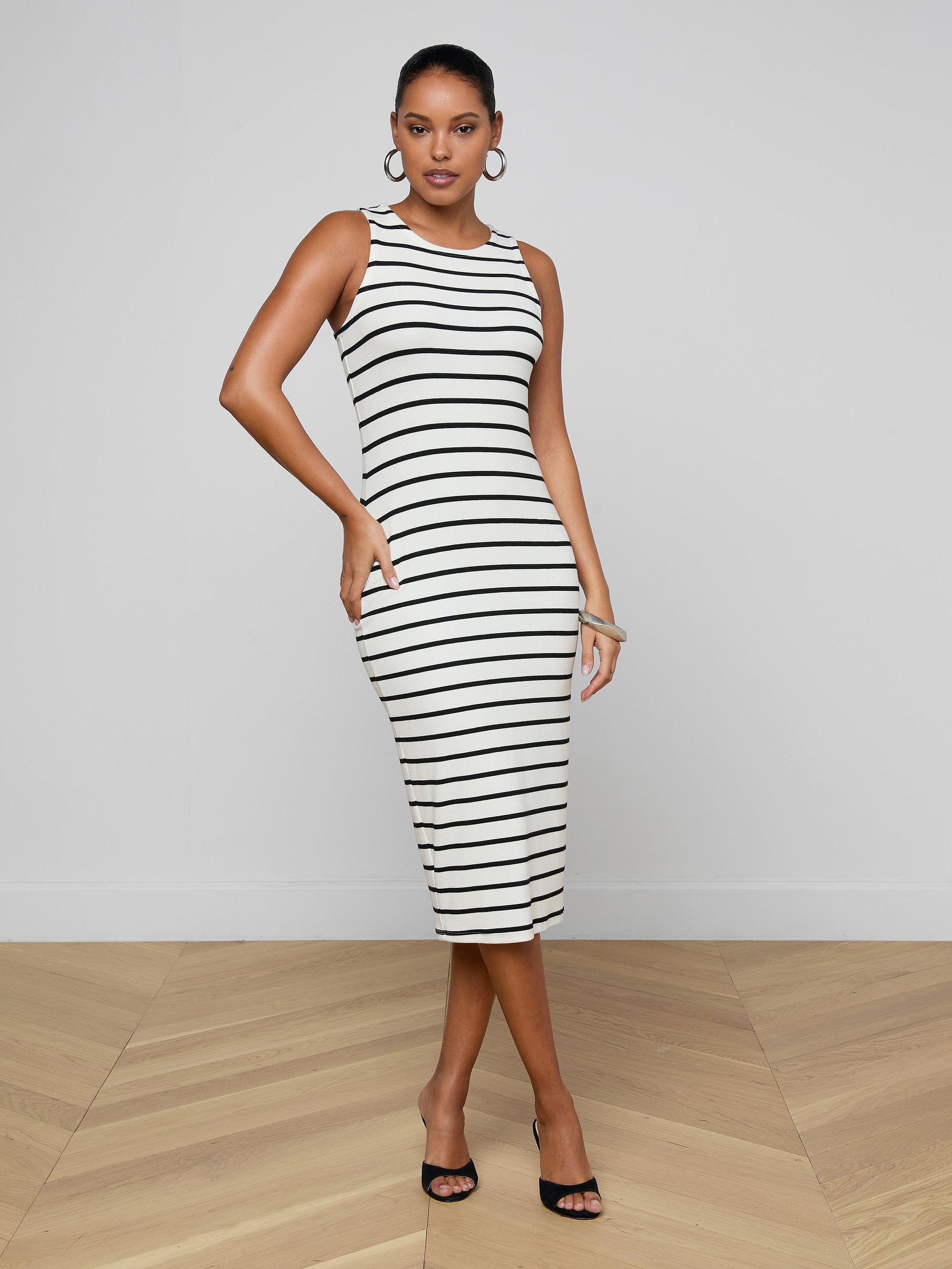 A woman wearing the Nura Racerback Midi Dress—an elegant sleeveless black and white striped jersey design featuring a racerback silhouette—pairs it with black heels and poses with one hand on her hip against a plain light background.