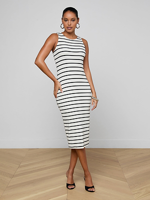 A woman poses against a plain light gray wall and wooden floor in the Nura Racerback Midi Dress, a sleeveless black-and-white striped day dress, paired with black heels, hoop earrings, and a bracelet.