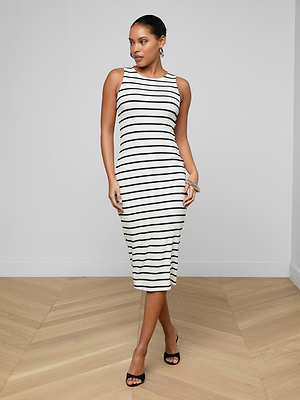 A woman poses indoors in the Nura Racerback Midi Dress—a knee-length, sleeveless black-and-white striped dress—paired with black heeled sandals, standing confidently on a light wood floor against a white wall.