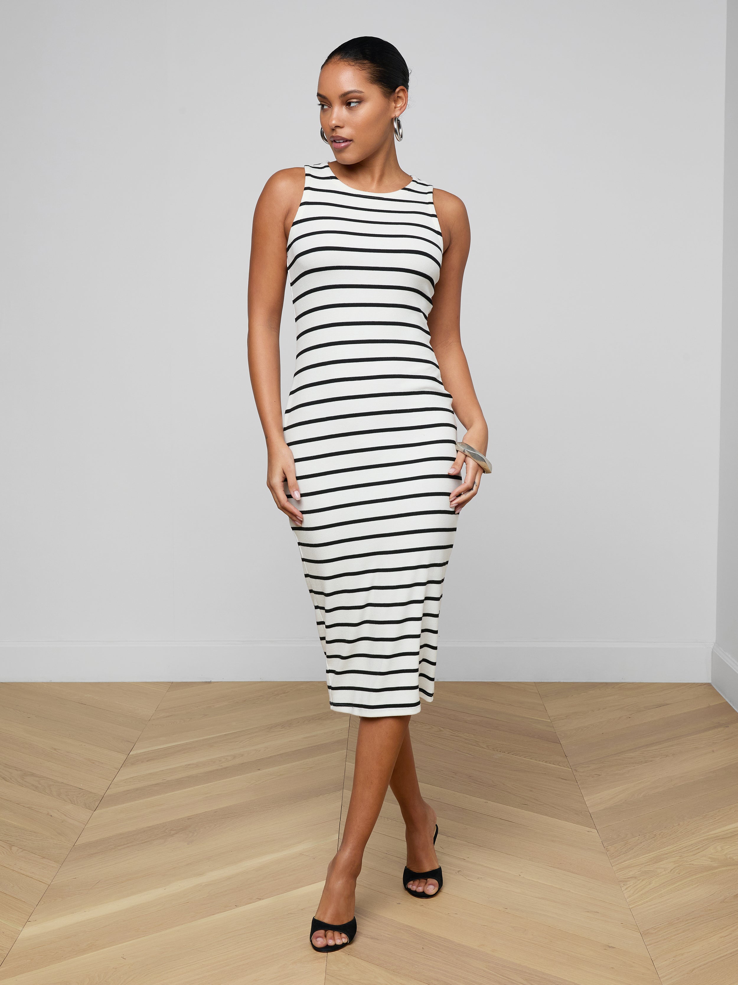 A woman stands against a plain white background wearing the Nura Racerback Midi Dress—a sleeveless, black and white striped jersey day dress with a racerback silhouette—paired with black heels.
