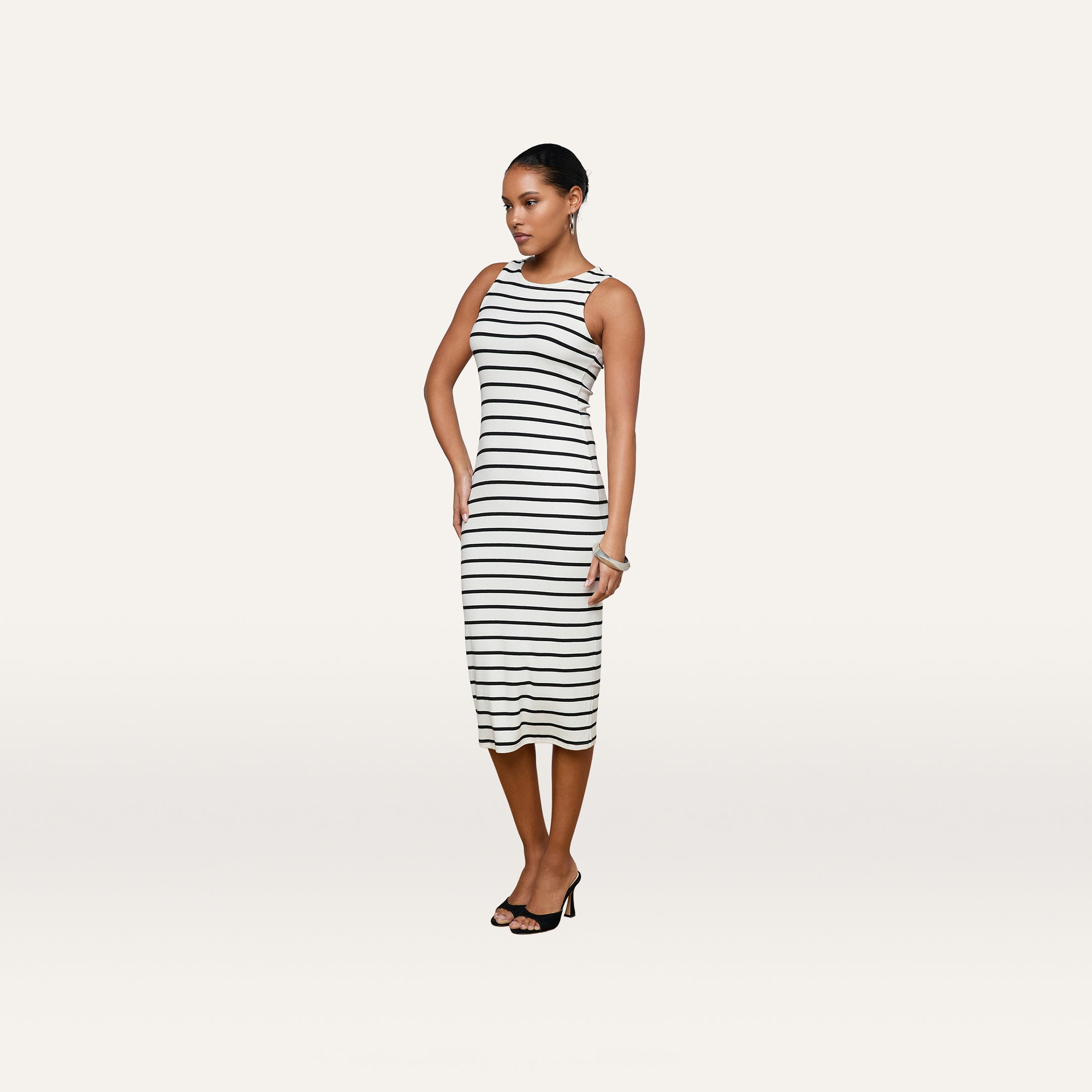 A woman is standing against a plain white background wearing the Nura Racerback Midi Dress—a sleeveless, black and white horizontal striped silhouette—paired with black heeled sandals.