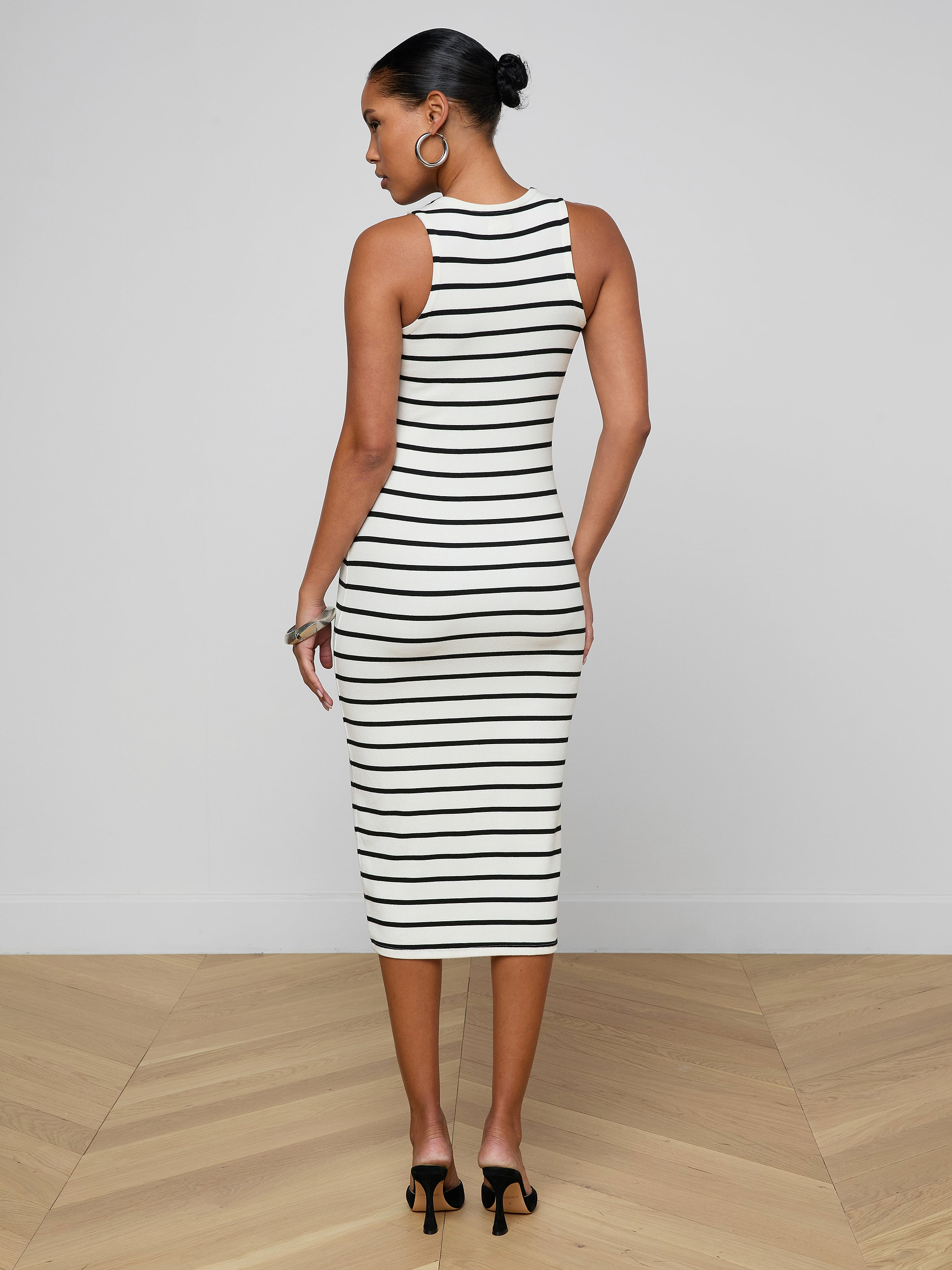 A woman wearing the Nura Racerback Midi Dress, a black-and-white striped, sleeveless, fitted midi dress with a racerback silhouette, stands facing away in black heels, her hair styled in a bun.
