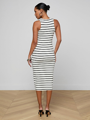 A woman stands on a wooden floor facing away, wearing the sleeveless Nura Racerback Midi Dress with black-and-white stripes, paired with black high-heeled sandals. Her hair is tied in a neat bun and she accessorizes with hoop earrings and bracelets.
