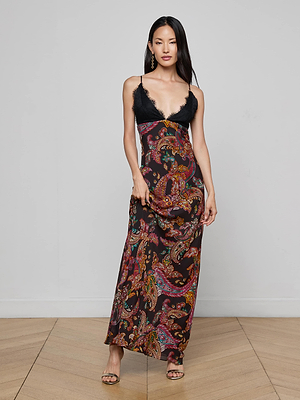 A woman poses gracefully indoors in the L'AGENCE Susanna Silk Lace Gown, which features a black lace top and a colorful paisley print skirt. She has long dark hair, wears earrings, and holds her dress with one hand.