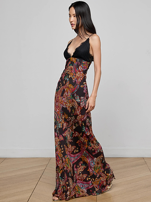 A woman with long, straight dark hair stands on a wooden floor wearing the L'AGENCE Susanna Silk Lace Gown—a long, flowing dress featuring a black bodice and colorful paisley print skirt. She faces to the side.