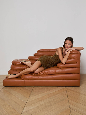 A woman in the L'AGENCE Inori Beaded Mini Dress reclines on a modern brown leather sofa with one arm on the backrest and one leg outstretched, set on a wood parquet floor against a white wall.