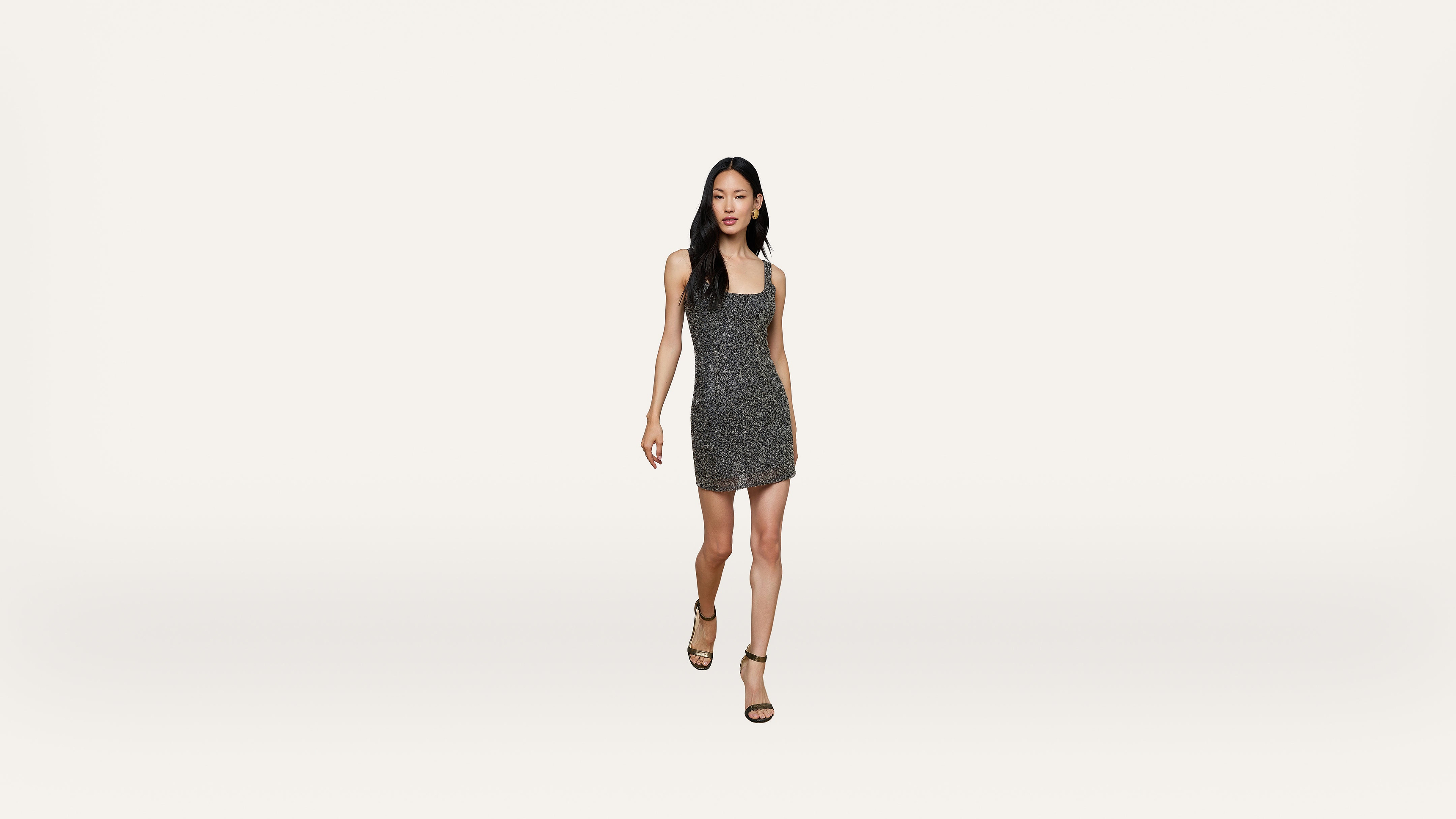 A woman wearing a sleeveless, fitted, sparkly gray dress and black heels walks confidently toward the camera against a plain white background.