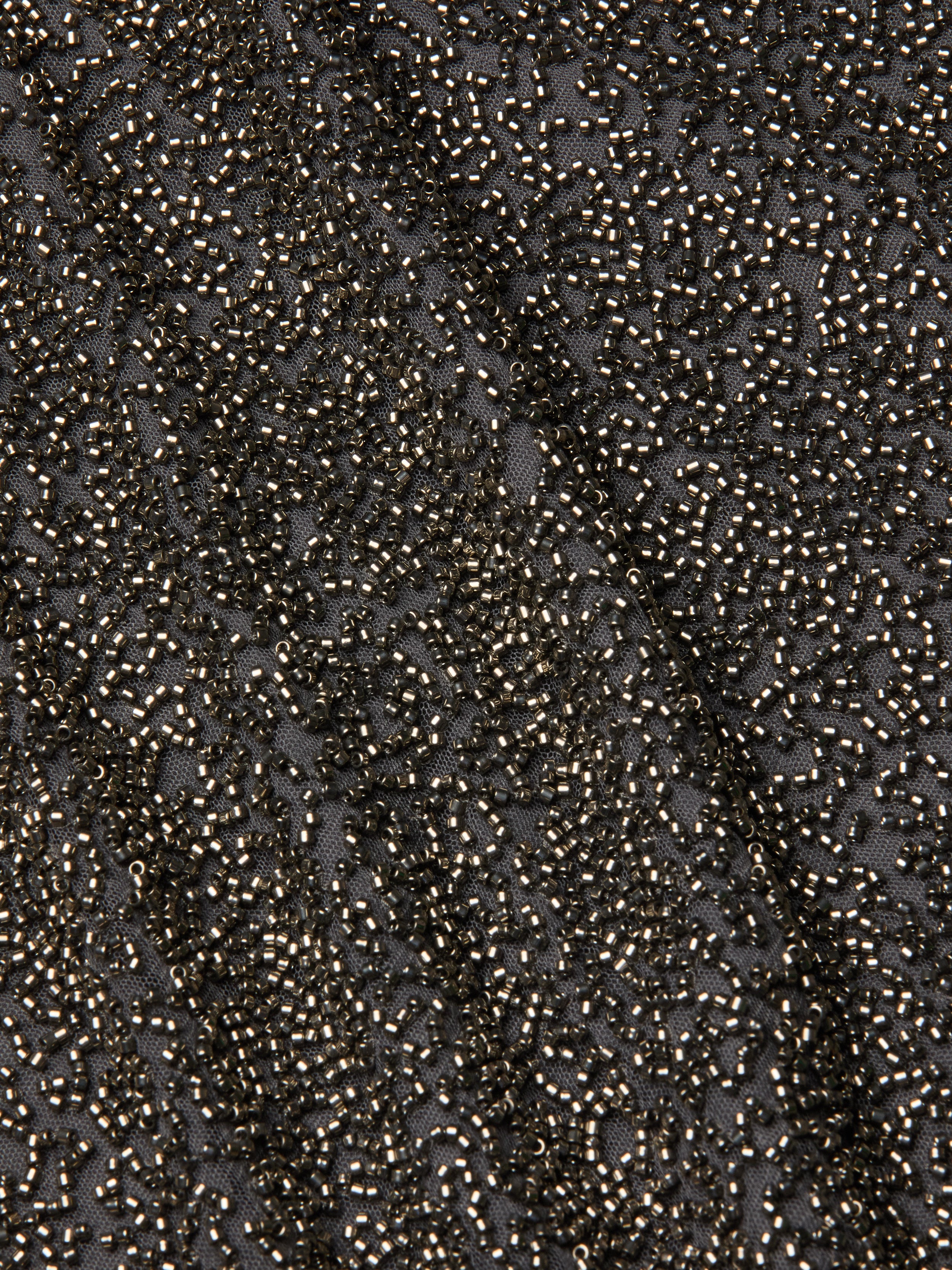 Close-up of the Inori Beaded Mini Dress, featuring a densely embroidered surface with metallic beads in black, creating a textured and shimmering effect; a diagonal seam is visible through the intricate pattern.
