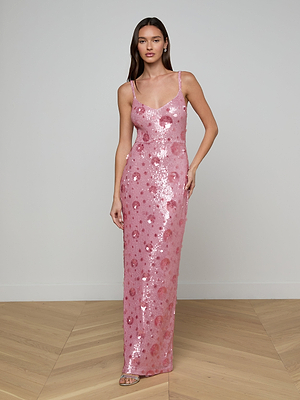 A woman with long brown hair and strappy sandals stands indoors on a wooden floor, wearing the Karma Sequin Maxi Dress adorned with pink sequins and a delicate floral pattern, against a plain light wall.