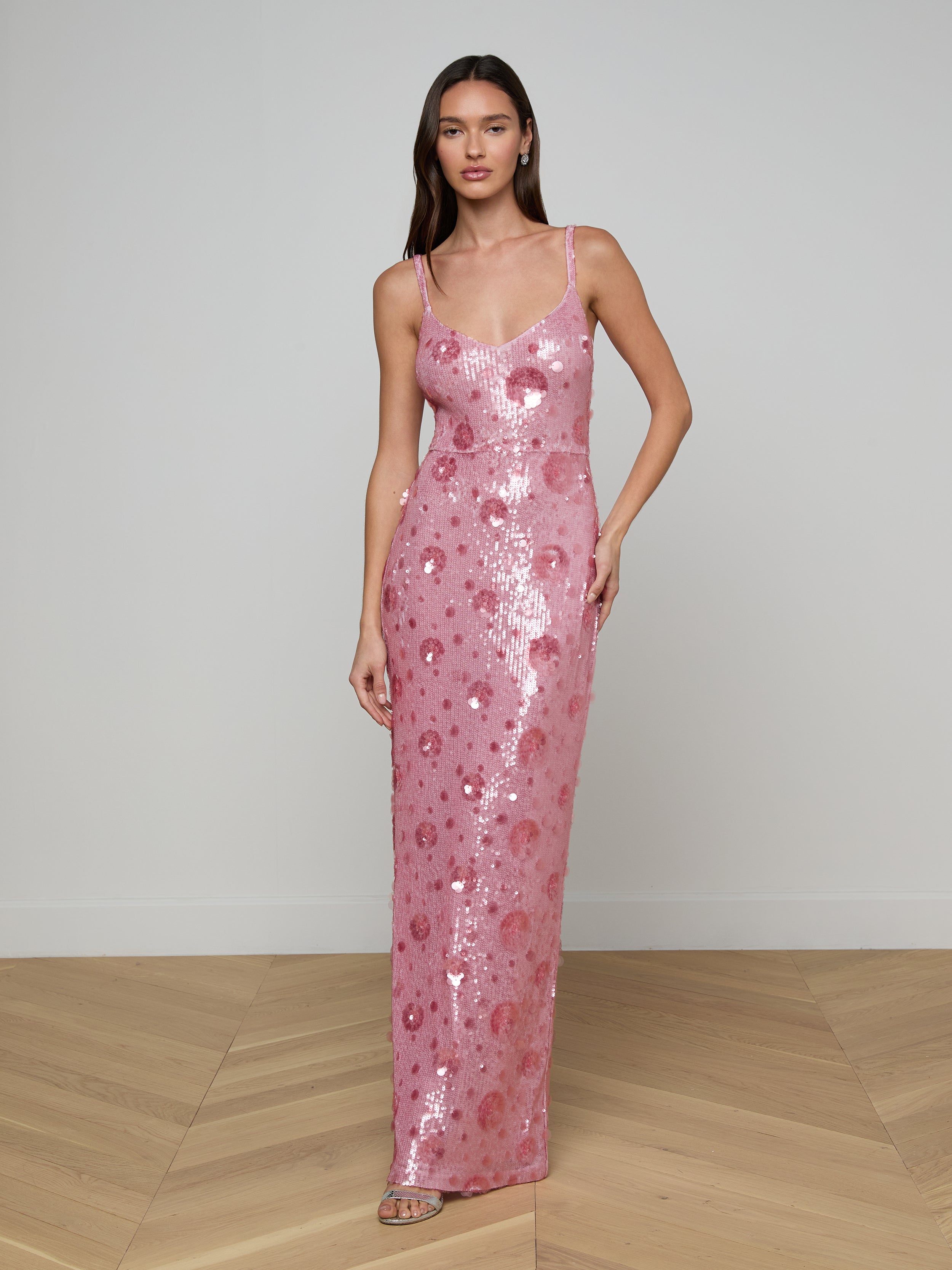 A woman wearing the Karma Sequin Maxi Dress, a long pink gown with thin straps, stands against a plain light background. She has straight hair and is posing with one hand on her hip.