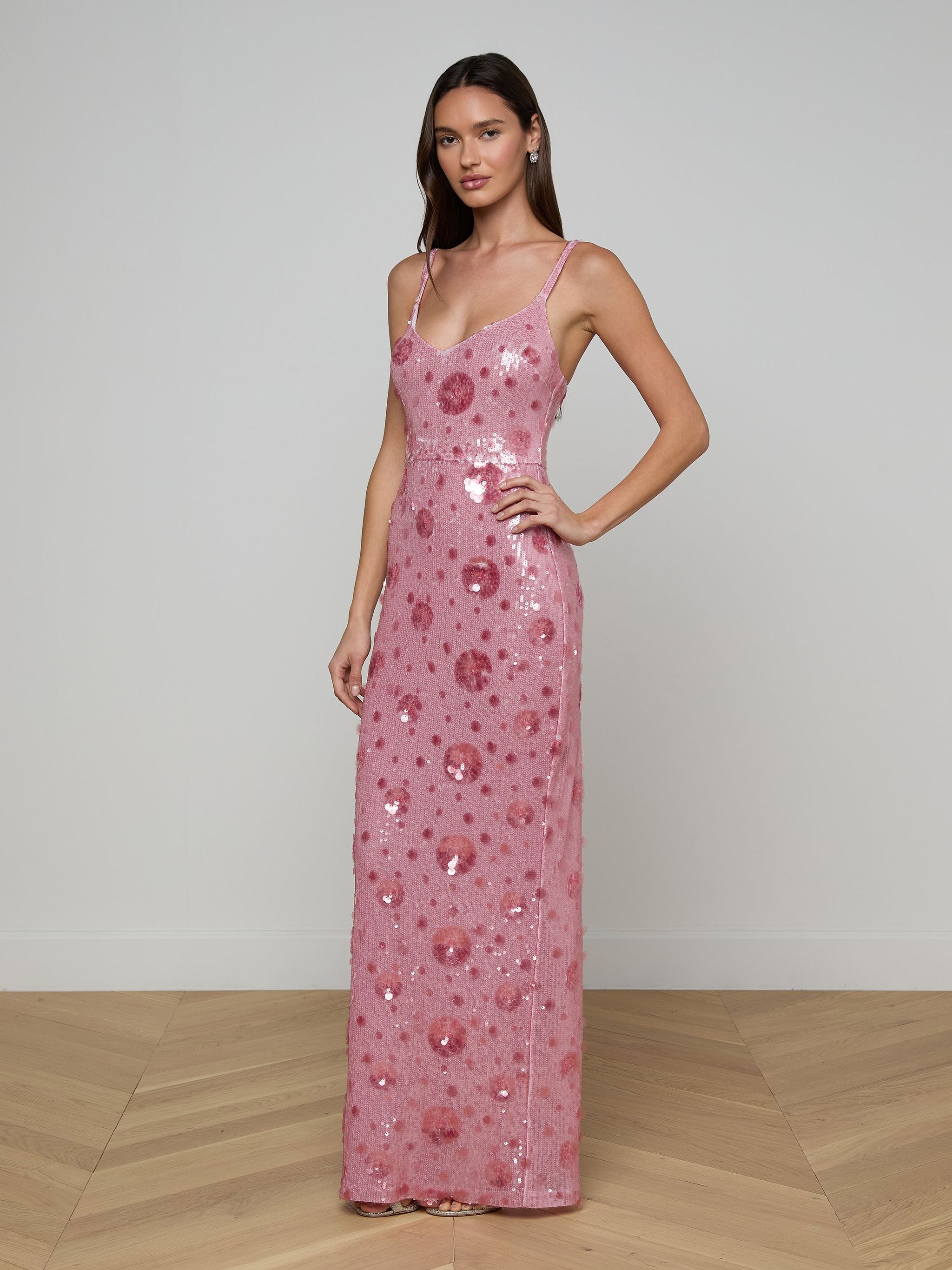 A woman wearing the Karma Sequin Maxi Dress—a sleeveless, floor-length gown with a fitted silhouette, spaghetti straps, and large pink sequin embellishments featuring subtle floral patterns—stands against a plain white background.