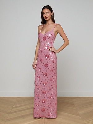 A woman poses against a white wall on a wooden floor, wearing the Karma Sequin Maxi Dress—a long, sleeveless pink dress with thin straps and a scoop neckline. She has long brown hair and rests one hand on her hip.