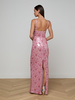 A woman faces away, wearing the Karma Sequin Maxi Dress in pink with thin straps and a back slit, paired with silver heels. She stands in a minimalist room with wooden floors and light-colored walls.
