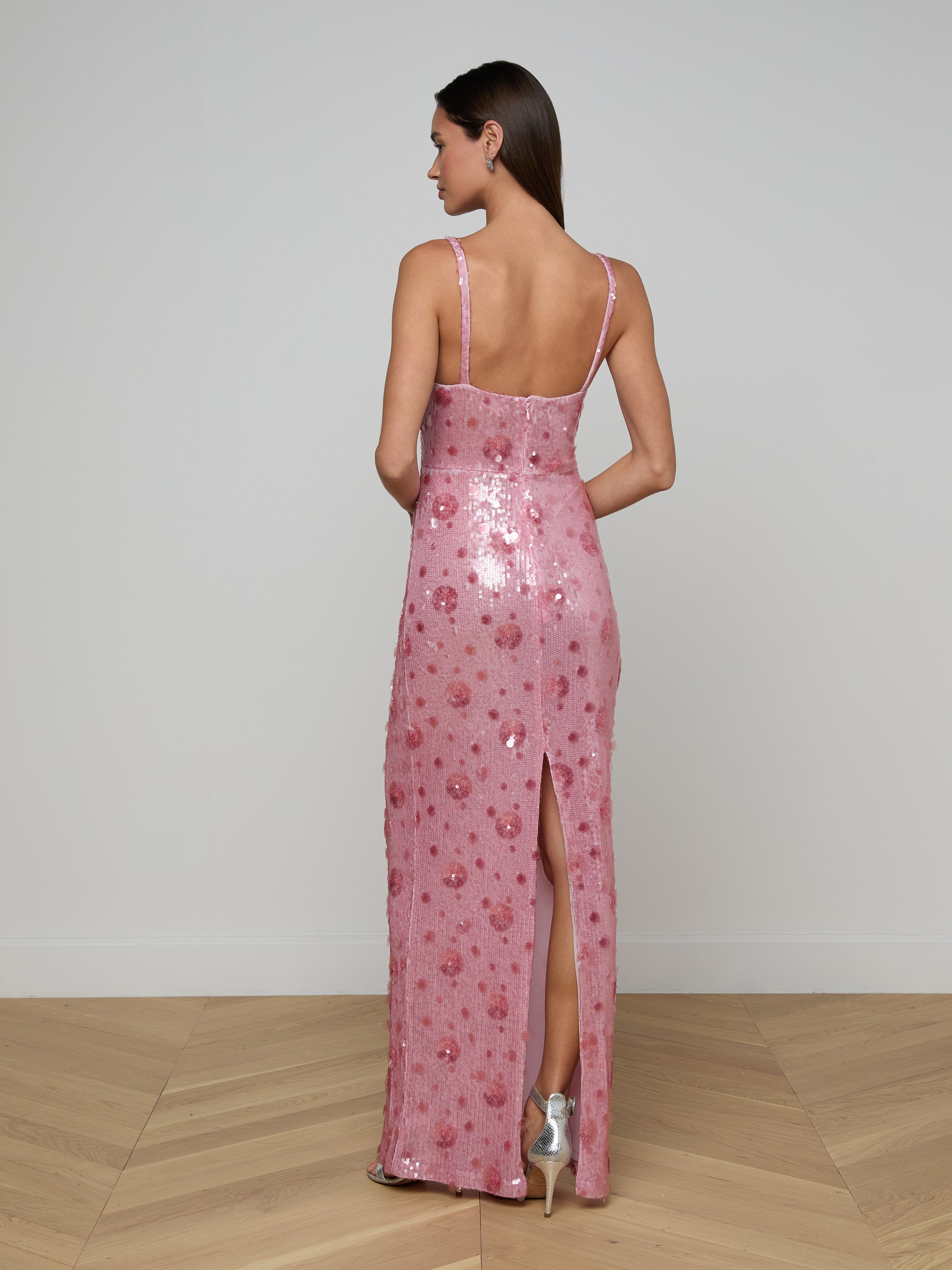A woman stands facing away, wearing the Karma Sequin Maxi Dress in pink, featuring thin straps and a back slit. She has straight brown hair and is wearing silver high heels. The background is plain white.