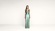 A woman stands in a minimal studio wearing a long, shimmering green dress with thin straps. She faces forward, hands by her sides, against a plain white background.