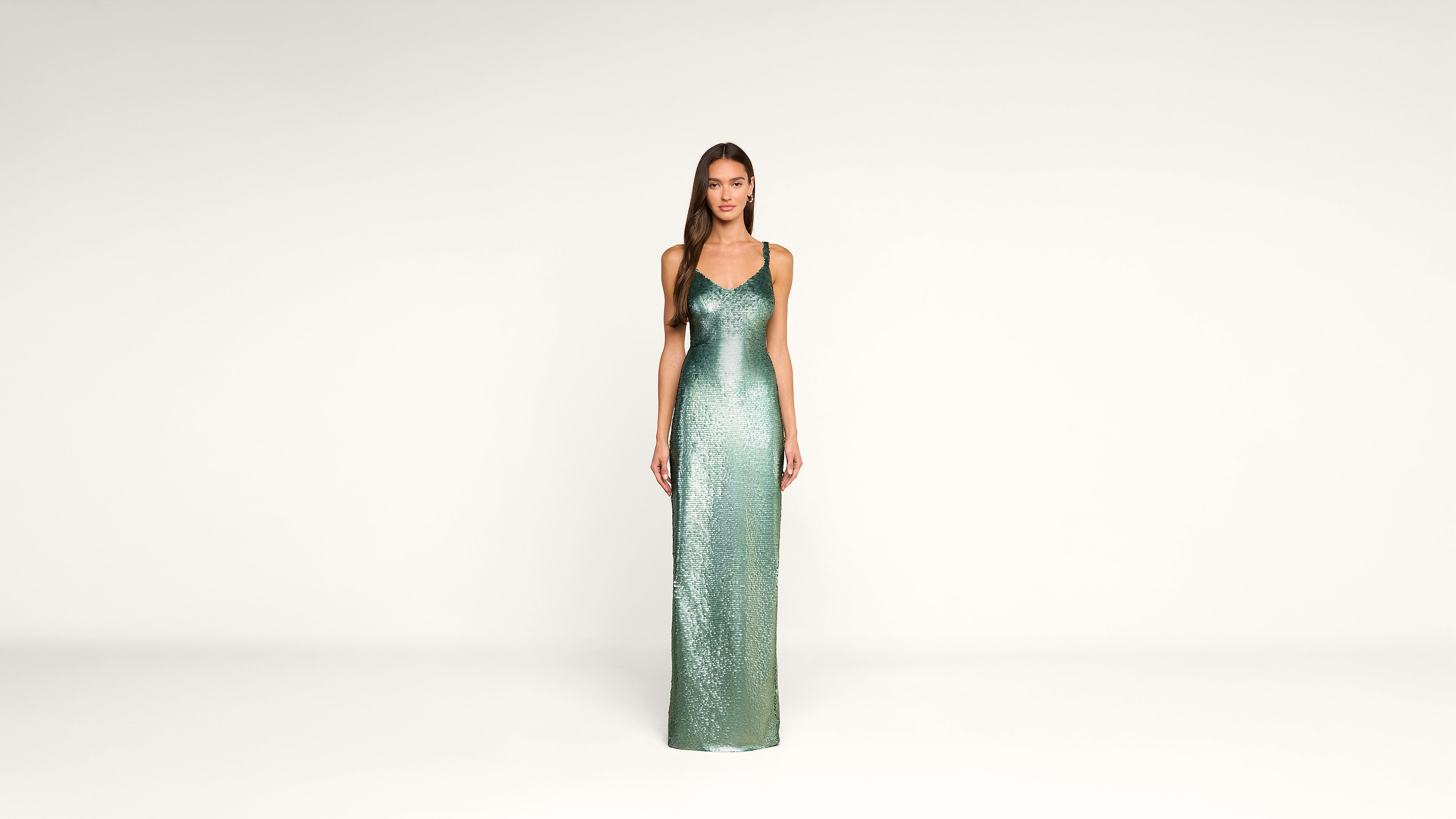 A woman stands in a minimal studio wearing a long, shimmering green dress with thin straps. She faces forward, hands by her sides, against a plain white background.