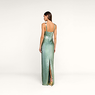 A woman stands facing away, wearing the Karma Sequin Maxi Dress—a fitted, sleeveless evening gown in shimmering green with a back slit—paired with gold heels. The background is plain and white.