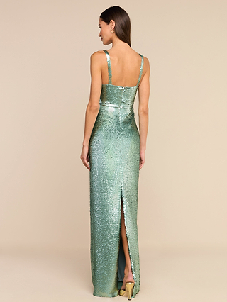 A woman stands facing away in a neutral studio setting, wearing the Karma Sequin Maxi Dress—a shiny, sleeveless green gown featuring a structured bodice and a high back slit.
