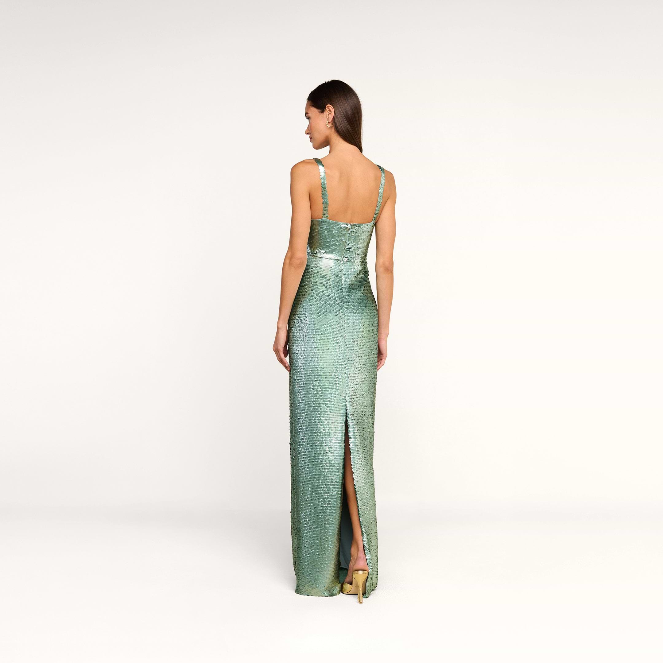 A woman stands facing away, wearing the Karma Sequin Maxi Dress—a fitted, sleeveless evening gown in shimmering green with a back slit—paired with gold heels. The background is plain and white.