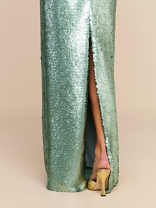 A person wearing the Karma Sequin Maxi Dress—a shiny, green sequin maxi dress with a high slit and structured bodice that reveals one leg—stands against a beige background, paired with gold high-heeled shoes.