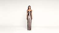 A woman stands against a plain white background wearing a long, shimmering, metallic gown with thin straps.