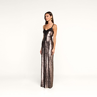 A woman stands against a plain white background, wearing the Karma Sequin Maxi Dress—a long, fitted gown with thin straps covered in sequins. She has straight brown hair, minimal jewelry, and a neutral expression.