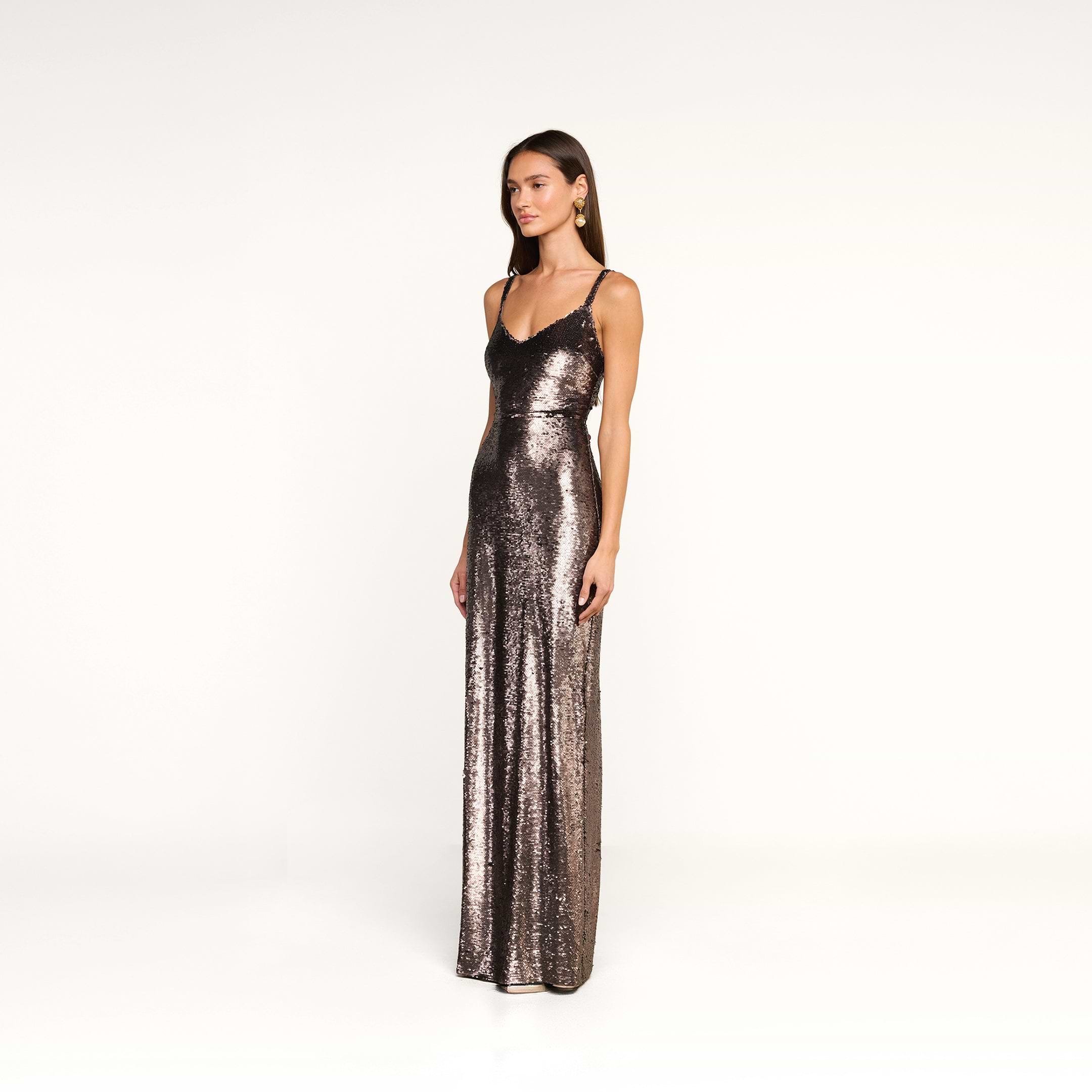 A woman stands against a plain white background, wearing the Karma Sequin Maxi Dress—a long, fitted gown with thin straps covered in sequins. She has straight brown hair, minimal jewelry, and a neutral expression.
