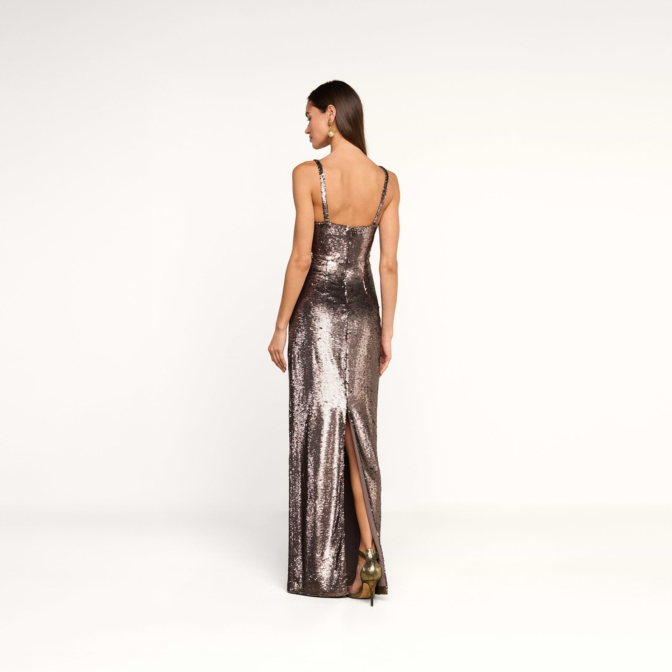A woman wearing the Karma Sequin Maxi Dress—a sleeveless, floor-length, fitted dress with sequins and a back slit—stands on a white background facing away from the camera.