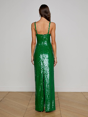 A woman faces away in the L'AGENCE Karma Sequin Maxi Dress, a fitted emerald green gown with thin straps, a structured sleeveless bodice, and a subtle back slit, flowing elegantly to the wood floor against a plain white wall.