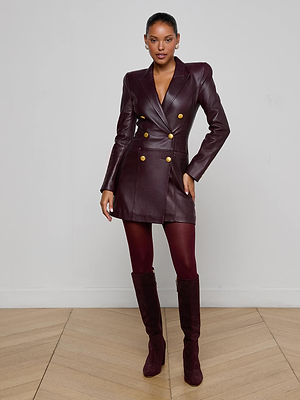 A woman stands indoors against a plain wall, wearing the Tora Leather Blazer Dress in burgundy with gold buttons, matching tights, and knee-high suede boots. Her hair is pulled back and one hand rests on her hip.