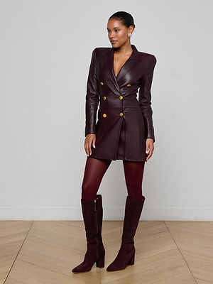A woman poses confidently indoors in the Tora Leather Blazer Dress—burgundy lambskin with gold buttons—paired with matching tights and knee-high suede boots against a white wall and wooden floor.