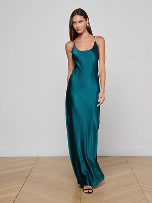 A woman with long brown hair stands on a wooden floor against a white wall, wearing the L'AGENCE Charmaine Silk Gown in teal with adjustable straps and black open-toe heels, looking slightly to the side.