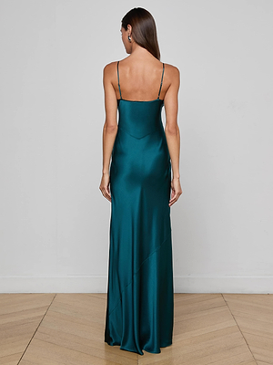 A woman with straight brown hair stands on a light wood floor against a plain wall, wearing the Charmaine Silk Gown by L'AGENCE—a long, teal bias-cut dress with adjustable straps that drapes elegantly to the floor.